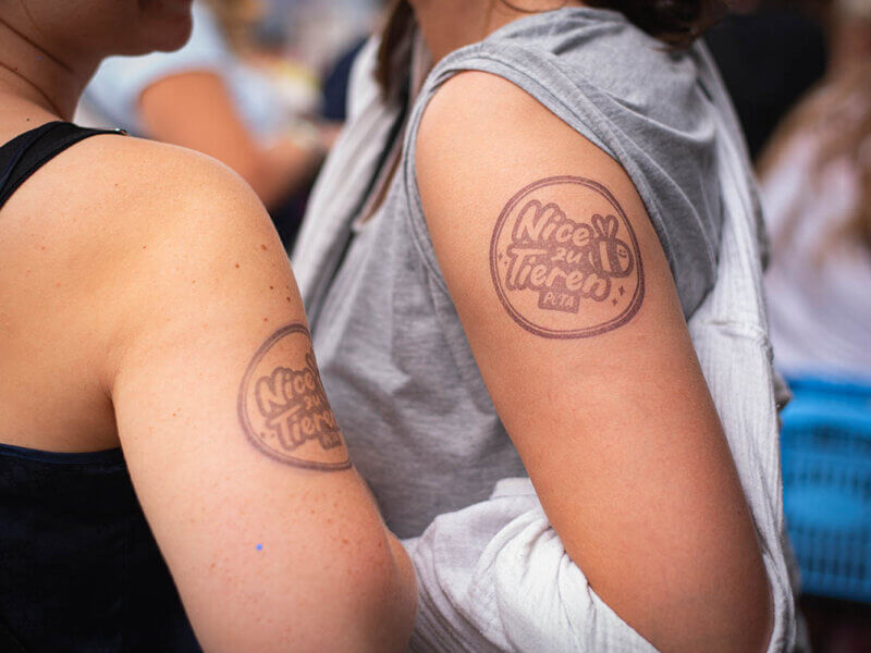 2 Frauen mit einem Stempel auf dem Arm mit der Aufschrift:"Nice zu Tieren PETA" und einer kleiner Biene.